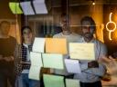 Drie mannen en een vrouw kijken in zakelijke setting naar post-it briefjes op een raam